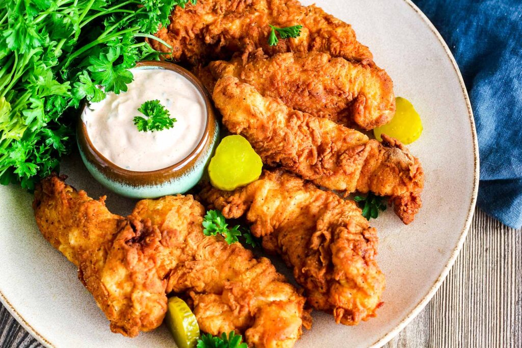 A plate of crispy chicken tenders with ranch, parsley, and pickles.