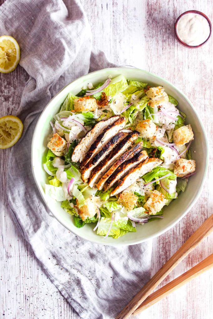 Bowl of blackened chicken salad with two lemon halves, a small bowl of dressing, and tongs.