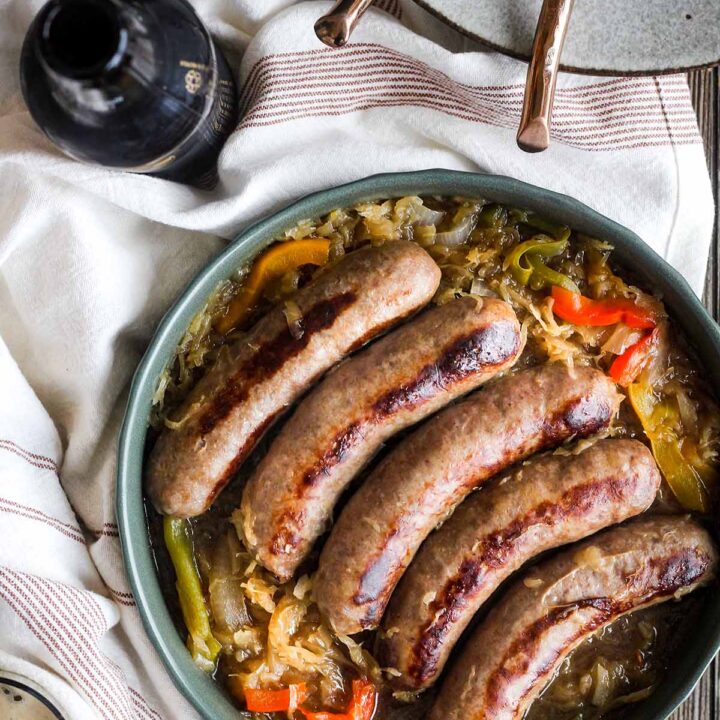 Bowl of brats with kraut with a bottle of Guiness, a cup, and a stack of plates with two forks.