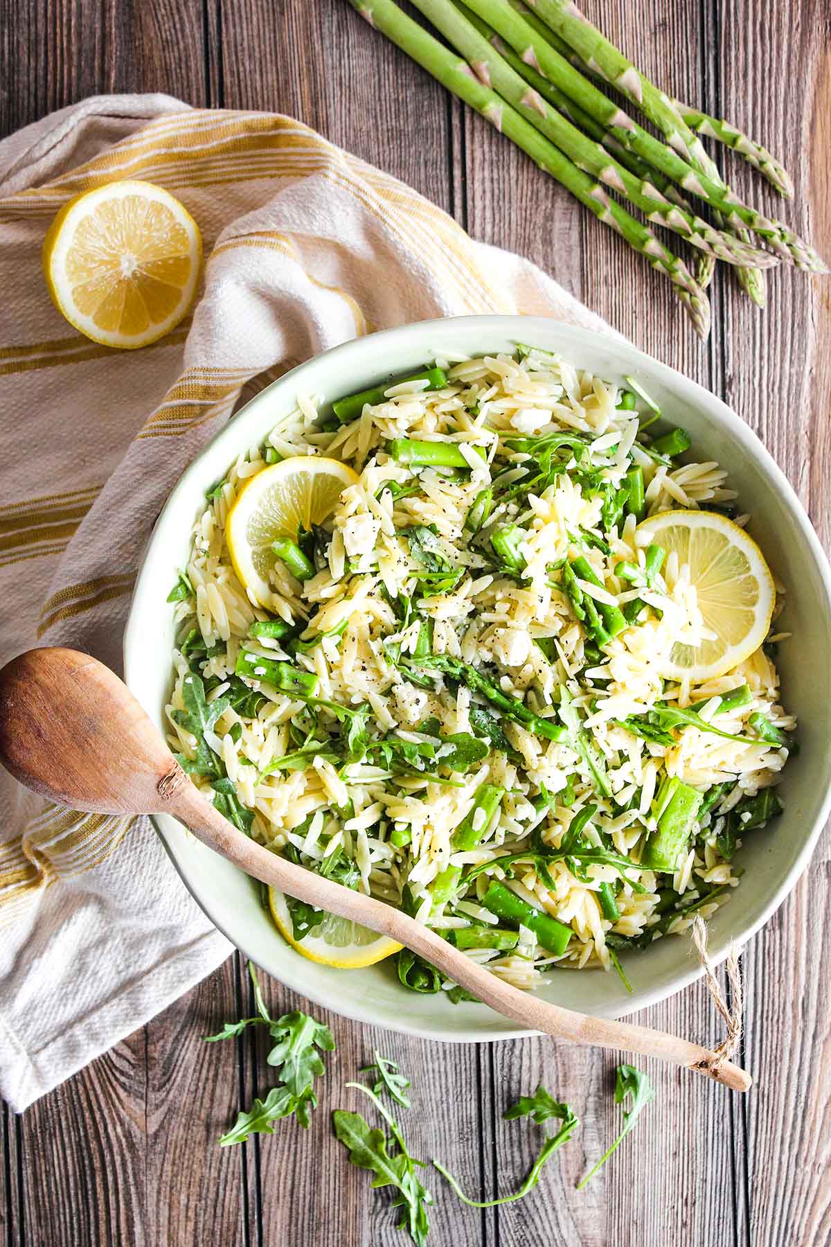 Bowl of orzo salad atop a kitchen towel with asparagus, lemon halves, and scattered arugula.