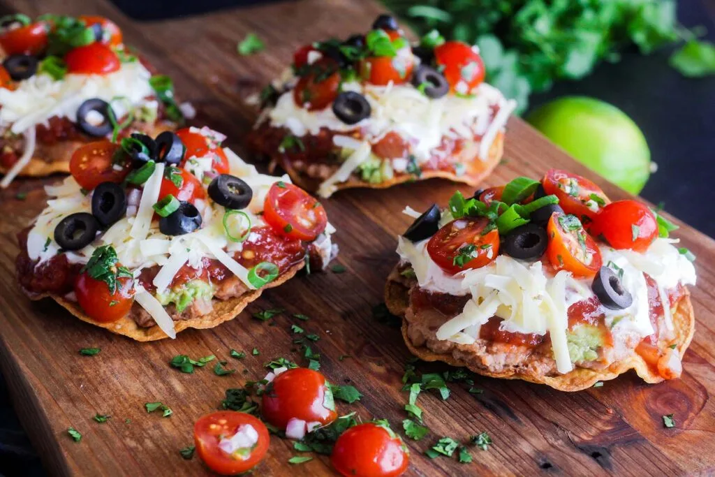 Four seven layer tostadas on a cutting board with tomatoes, cilantro, and a lime.