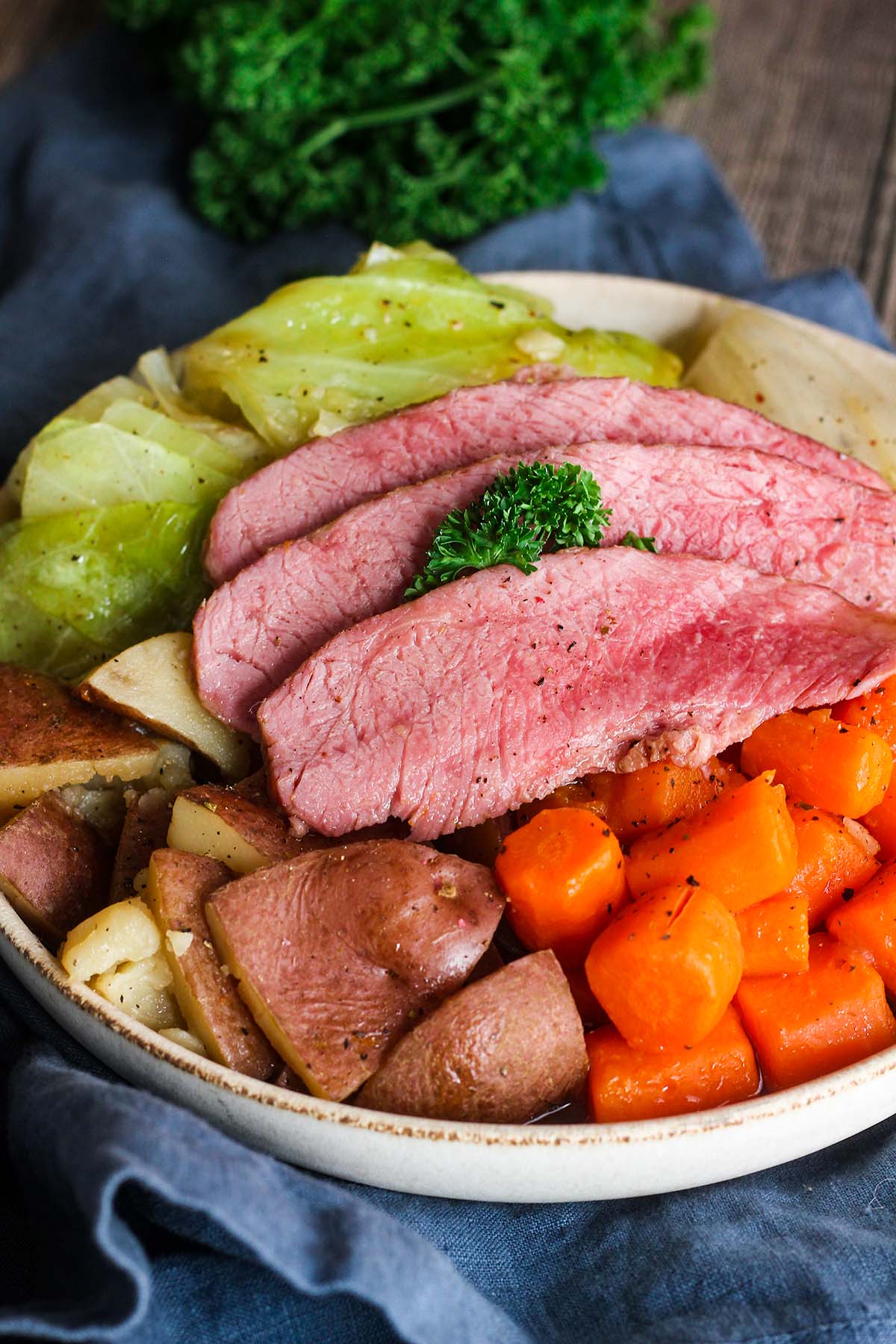 Three slices of corned beef atop cooked veggies in a bowl with a bunch of parsley and a blue kitchen towel.