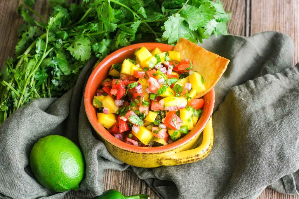 A bowl of mango salsa with a chip on top of a towel with cilantro and a lime.