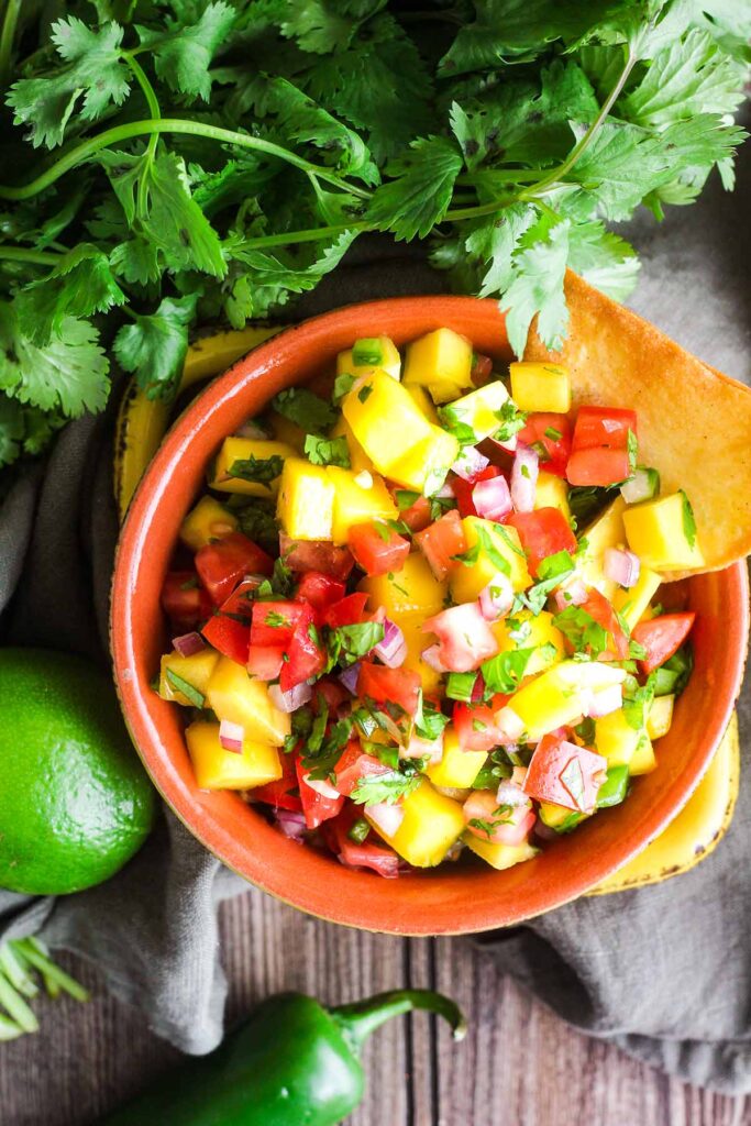 Bowl of fruit salsa next to a bunch of cilantro, a lime, and a jalapeno.