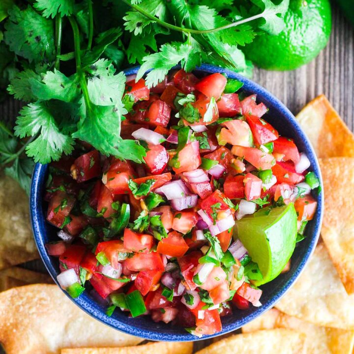 Bowl of pico de gallo surrounded by chips, a bunch of cilantro, a lime, and jalapeno.