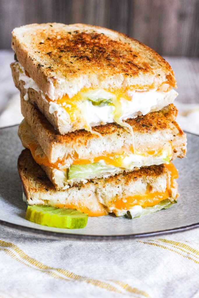 Four halves of pickled grilled cheese sandwich stacked on a plate with a sliced pickled.