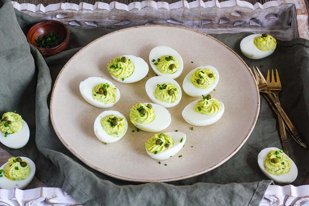 Plate of deviled eggs atop a serving tray with a dish towel, appetizer forks, and a small bowl of chives.