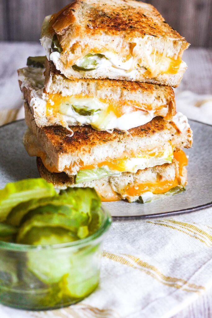 Stack of grilled cheese with pickle sandwiches on a plate with a small bowl of pickles.