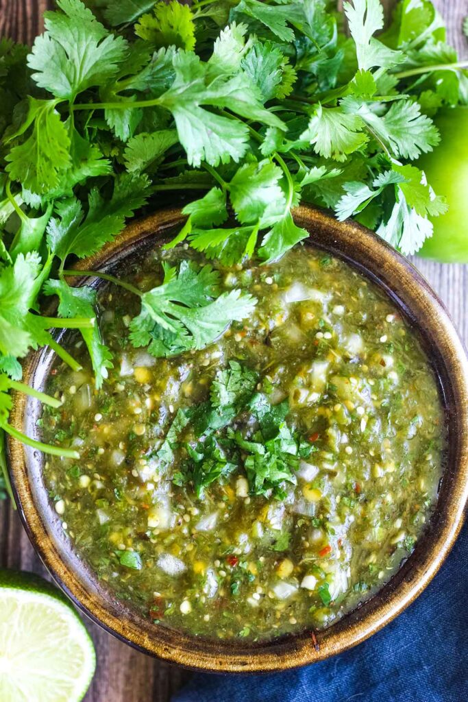 Bowl of roasted salsa verde surrounded by cilantro, a tomatillo, and a lime halve.