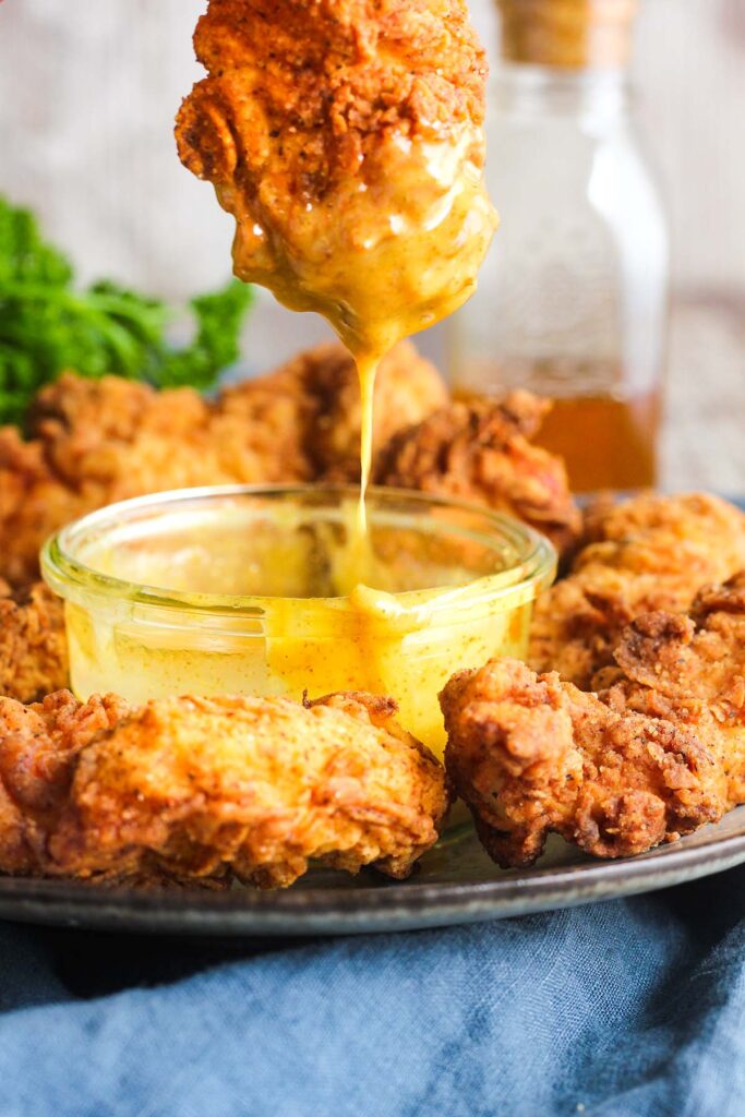 Chicken strip dripping with honey mustard atop bowl and plate of chicken.