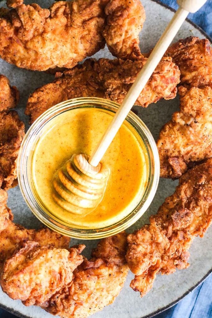 Close up of bowl of honey with honey stick surrounded by crunch chicken tenders.