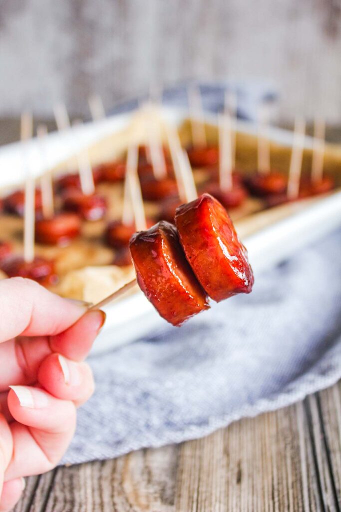Hand holding a toothpick with two kielbasa bites with a tray in background.