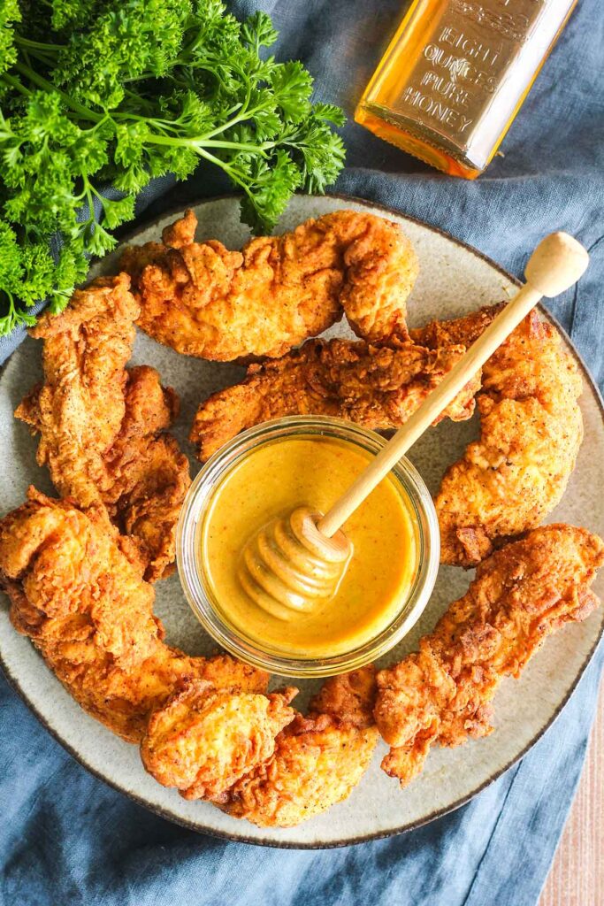Plate of chicken tenders with a bowl of honey mustard in center surrounded by jar of honey and bunch of parsley.