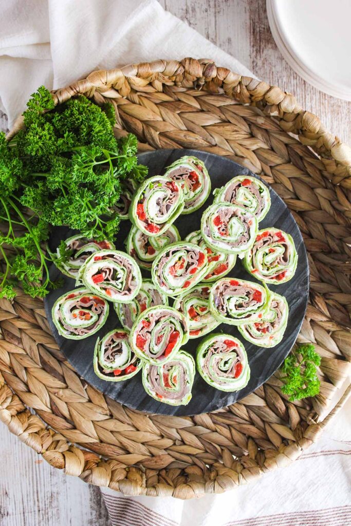 Tray of boursin pin wheels surrounded by parsley with a stack of plate and a dish towel.