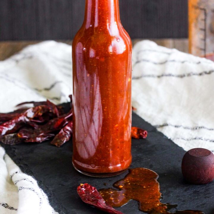 An unlidded bottle of chile de árbol hot sauce on a tray with spilled sauce and scattered chiles.