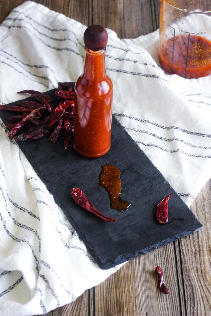 Bottle of hot sauce on a tray with spilt sauce and scattered dried chiles with a cup of sauce in background.