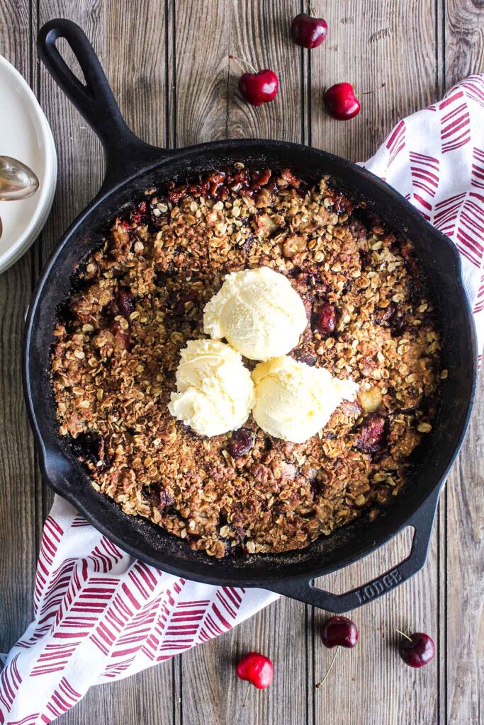 Finished crisp in a cast iron skillet topped with three scopes of ice cream atop a kitchen towel with scattered fresh cherries.
