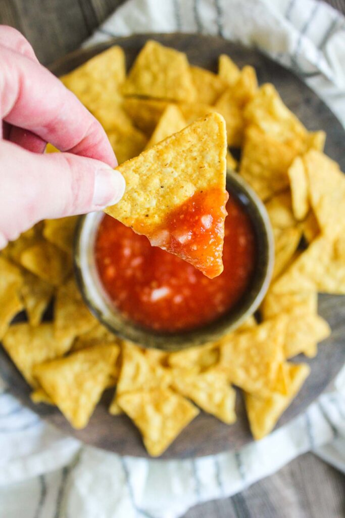 Hand holding chip dipped in salsa above platter with chips and salsa.