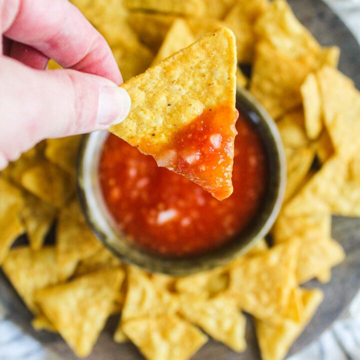 Hand holding chip dipped in salsa above platter with chips and salsa.