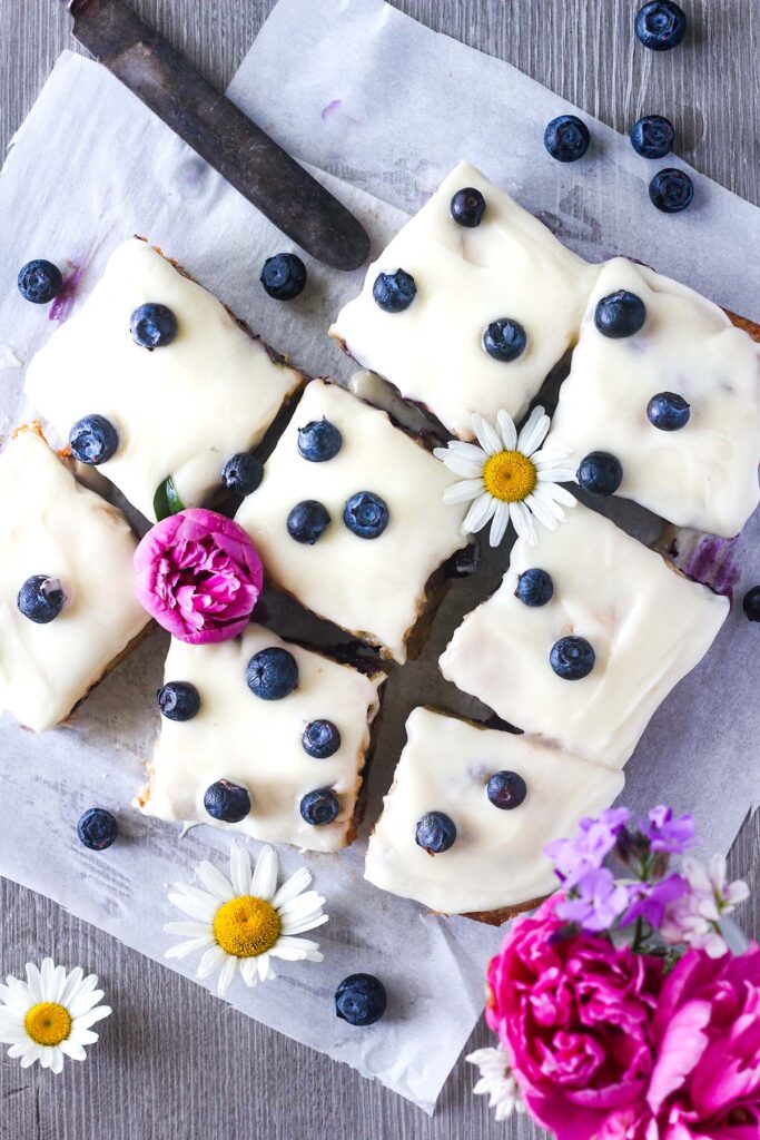 Sliced blueberry blondies topped with frosting and blueberries with scattered flowers.